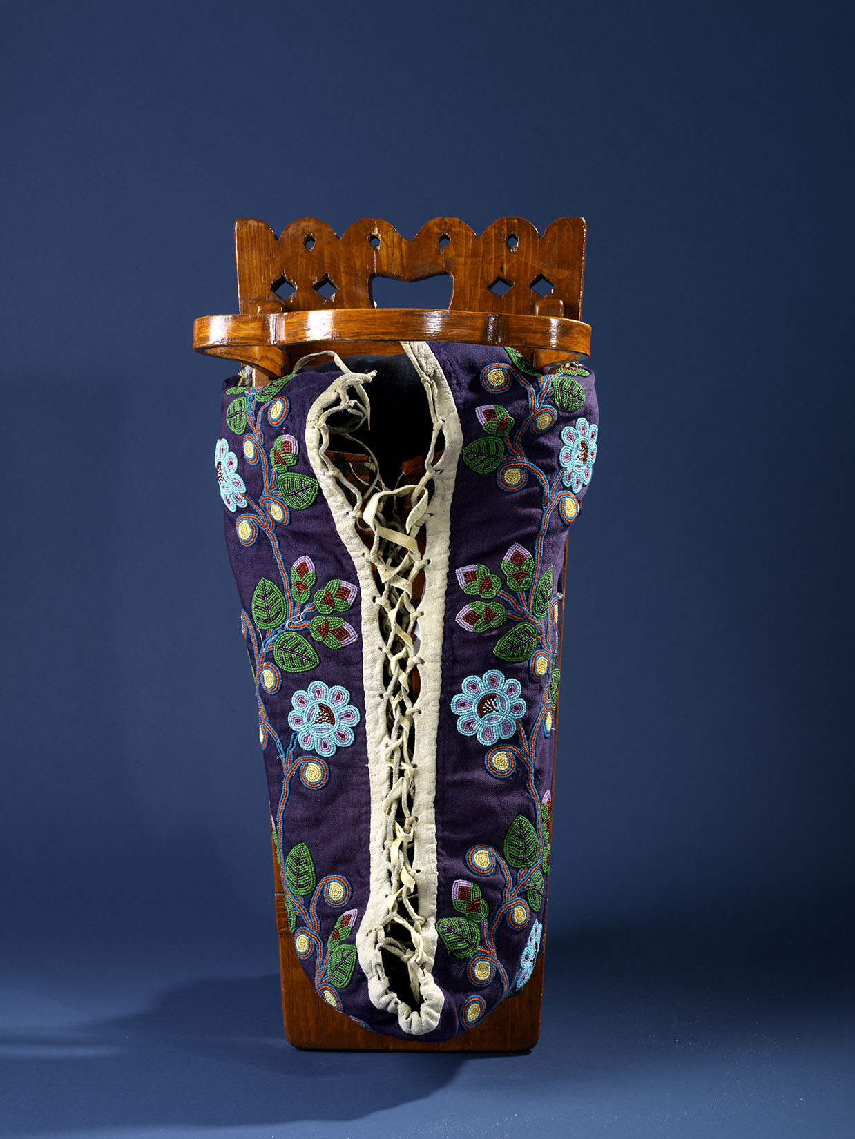 Cradleboard and moss bag with a varnished wood frame. The fabric of the moss bag where the baby is held is a deep purple-blue, decorated with a beaded pattern of light blue, purple and green flowers and blossoms. Other materials are wood, cotton cloth, glass beads, metal
