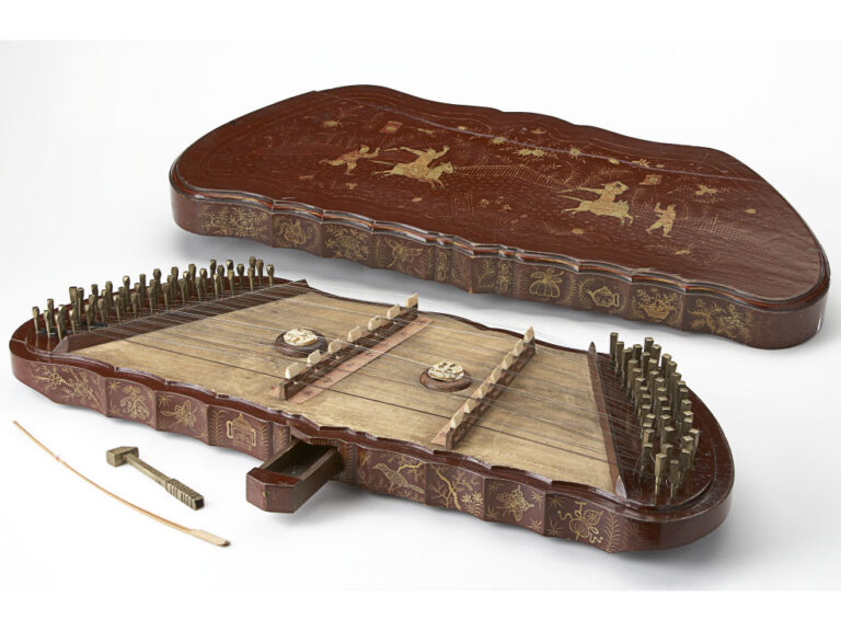 A wood engraved dulcimer string instrument. Tuning dials line either side of the instrument with a small drawer in the front of it
