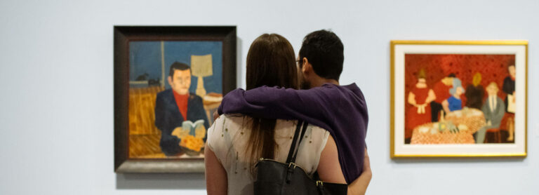Two people hug while looking at paintings at Glenbow. On the left is a painting of a man reading a book; on the right is a family gathering around a table